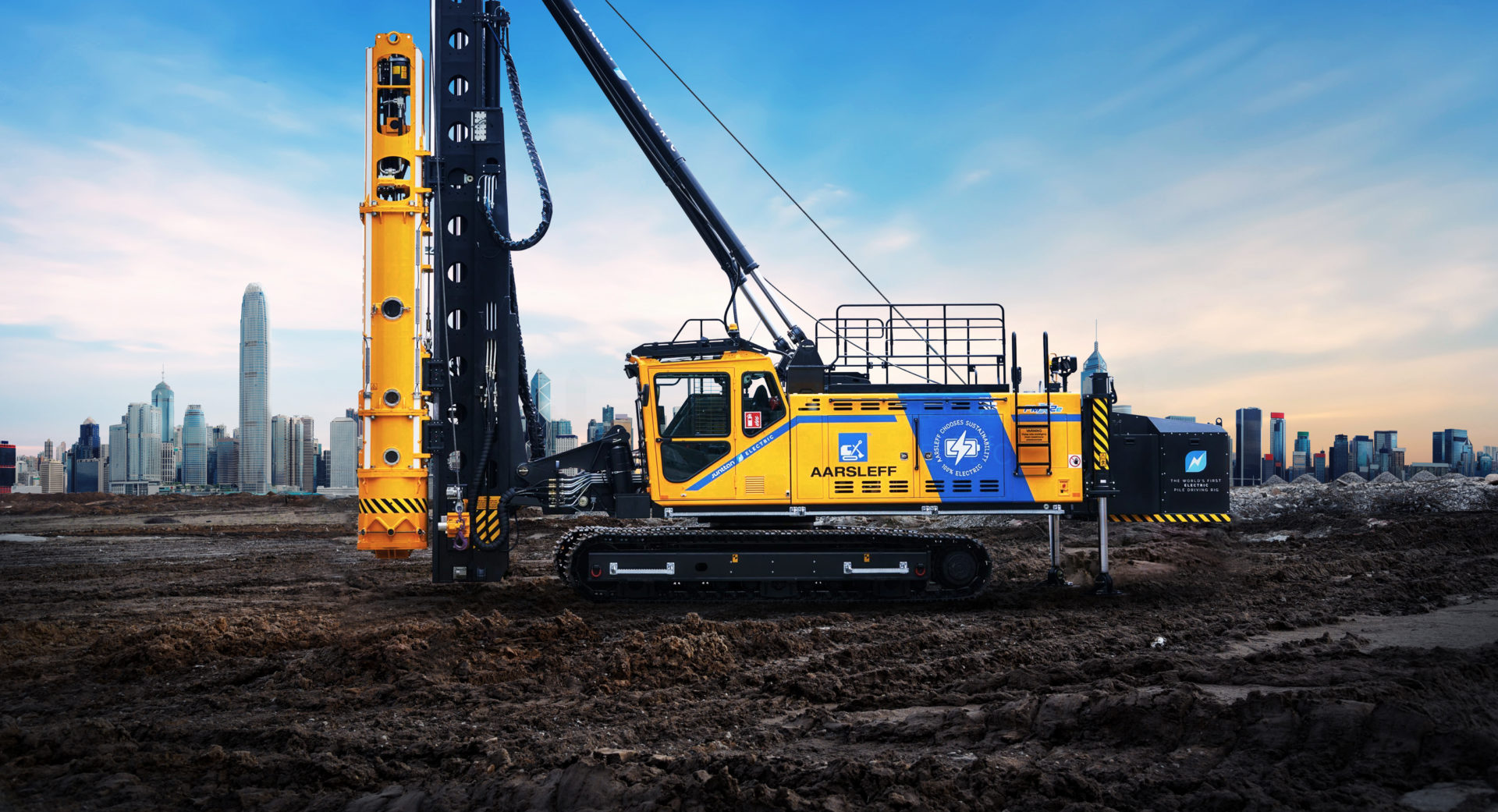 A heavy-duty blue and yellow pile driving rig, featuring Kempower EV charging solutions, positioned on an expanse of undeveloped land with a city skyline in North America in the background under a dramatic
