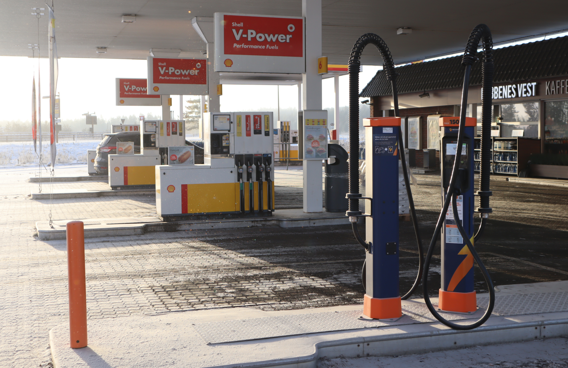 An empty petrol station on a sunny day with light snow cover, advertising Kempower EV charging solutions, with a small café sign in the background.