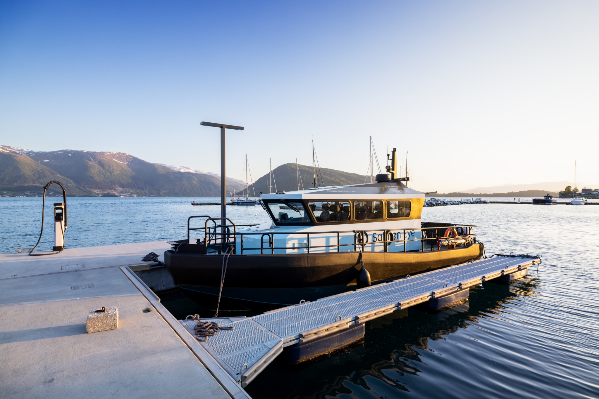 A boat docked at a pier by a calm body of water during sunset, with mountains in the background and a clear sky overhead.