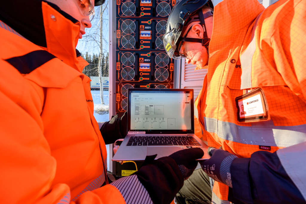 Two professionals wearing high-visibility jackets and safety helmets discussing data on a laptop at an industrial site.