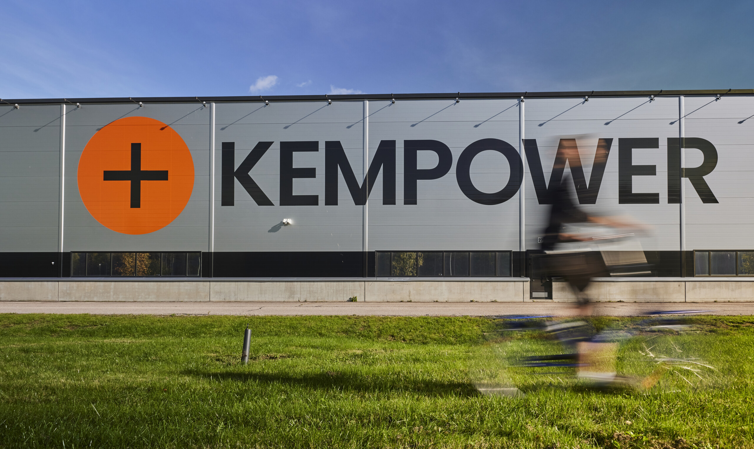 A person rides a bike past a large gray building with an orange plus sign and the word "KEMPOWER" in bold black letters.